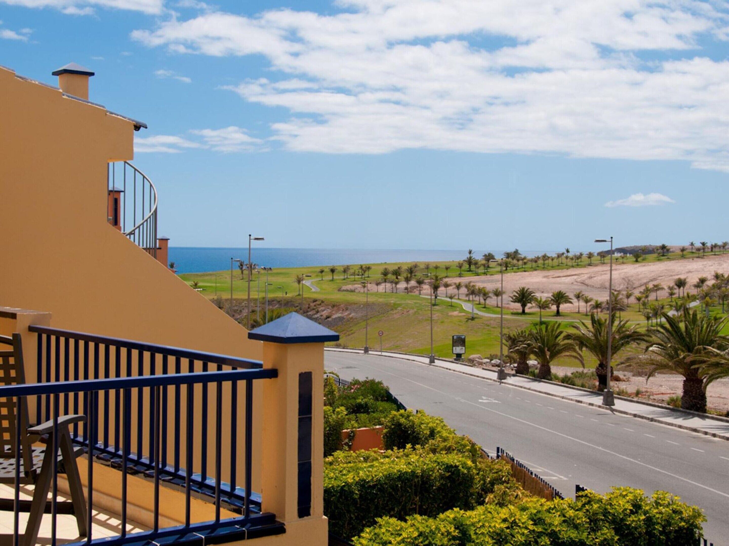A residential building with a balcony overlooking a coastal road with palm trees and a view of the sea.