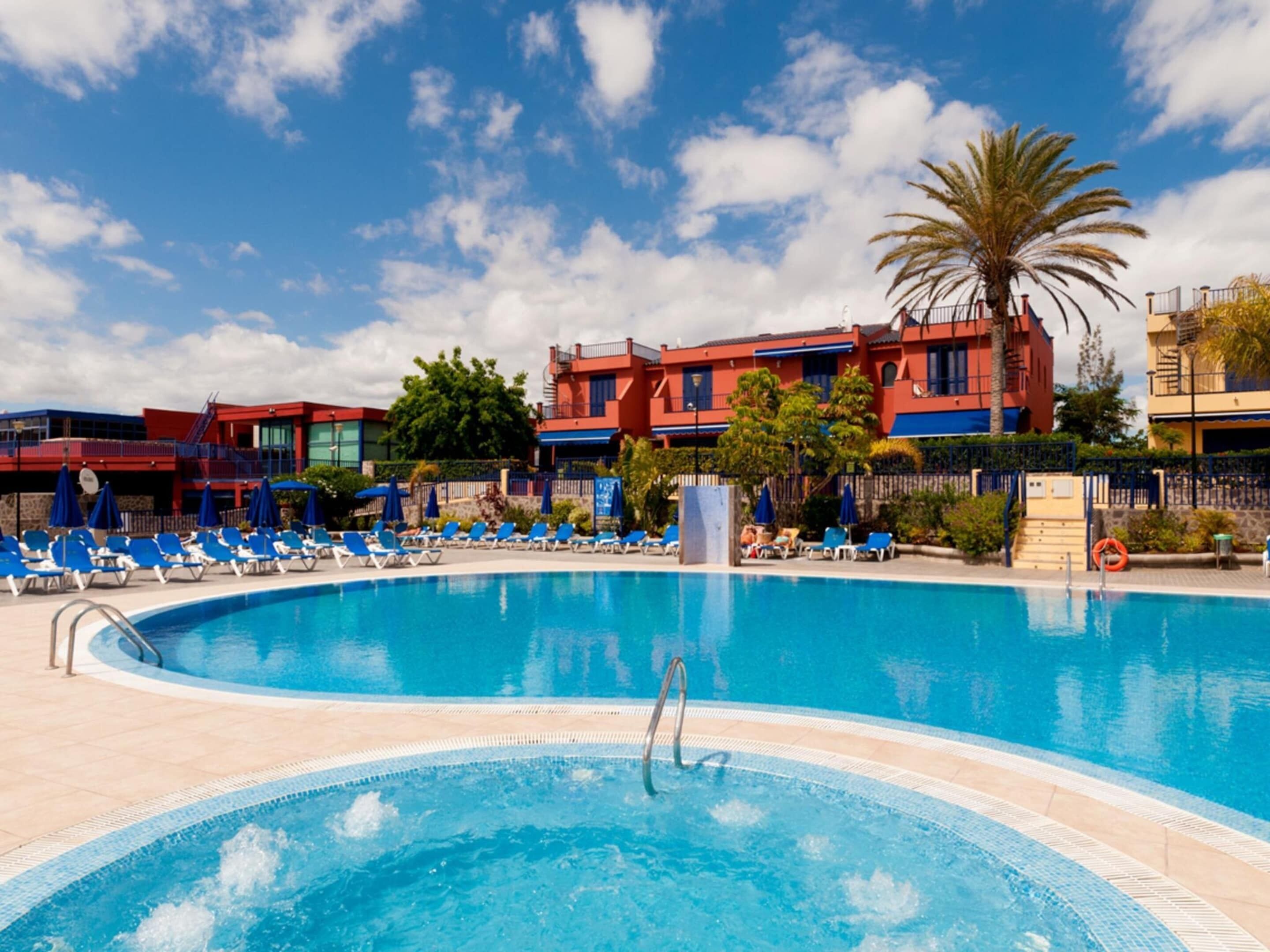 A swimming pool with lounge chairs and a palm tree, surrounded by buildings with red and blue facades.