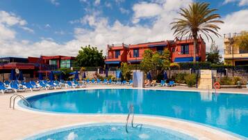 A swimming pool with lounge chairs and a palm tree, surrounded by buildings with red and blue facades.
