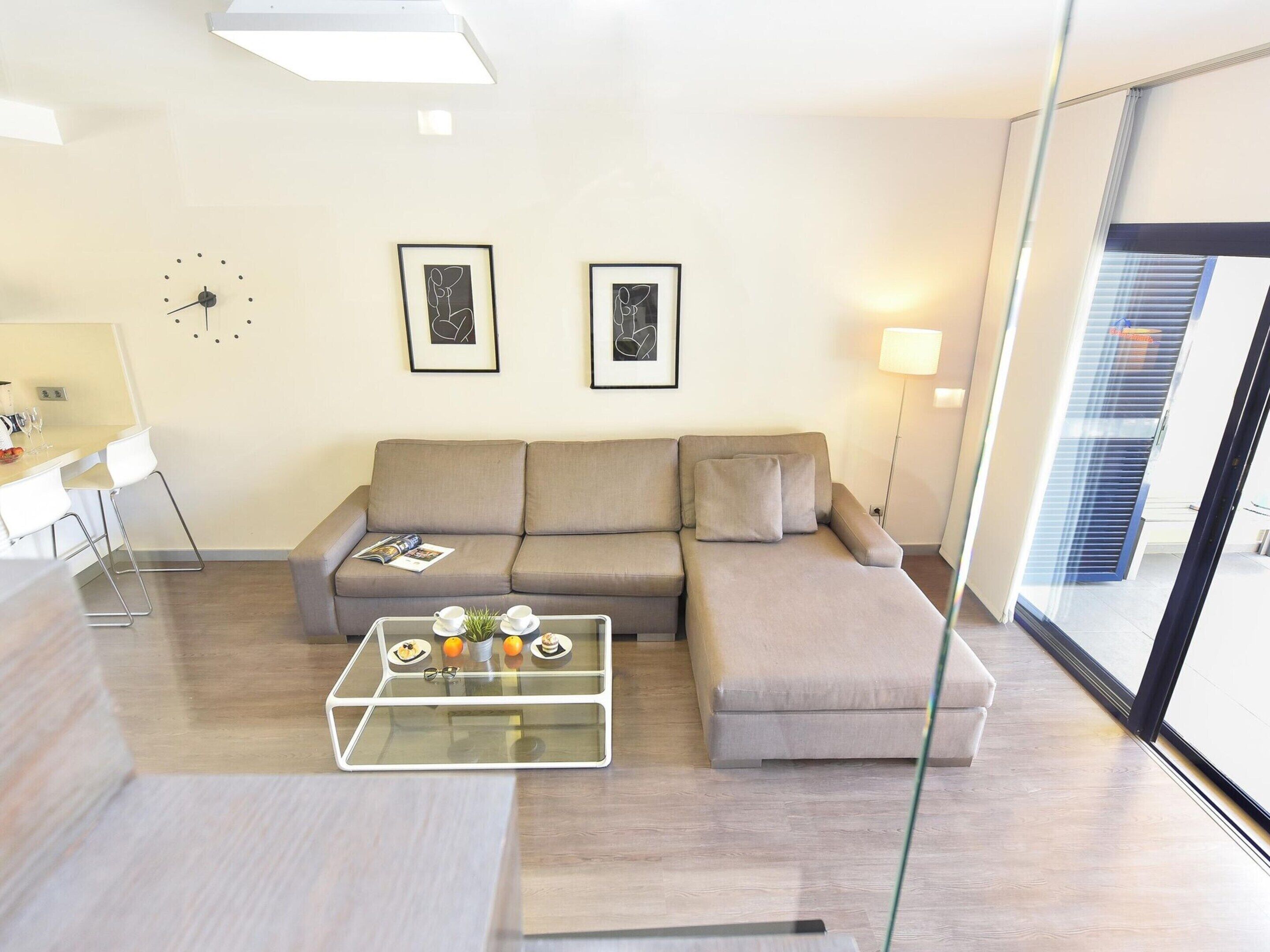 A modern living room with a sofa, a glass coffee table, and a dining area with chairs. There are two framed pictures on the wall, a clock, and a floor lamp.
