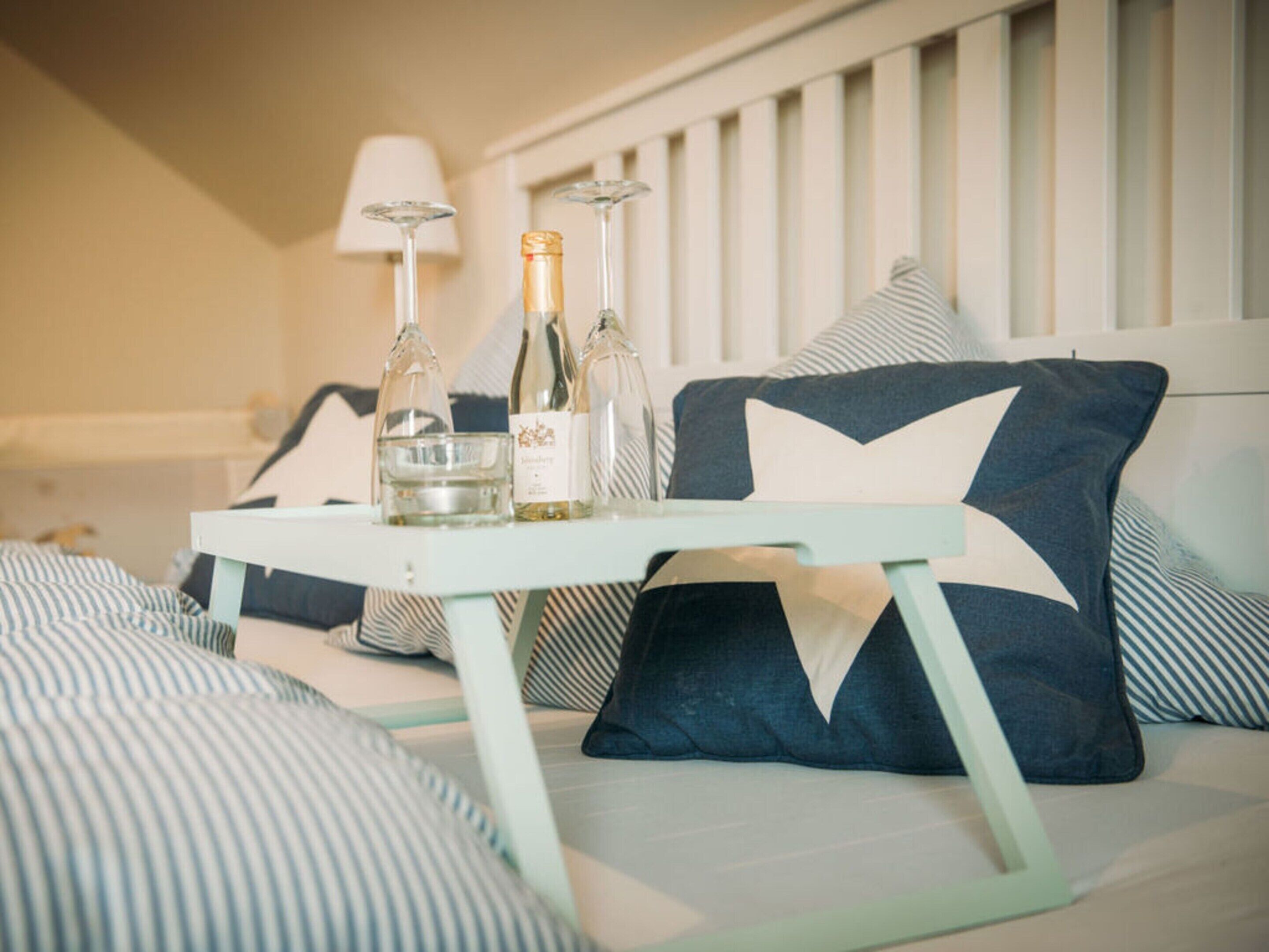 Een bed met een wit hoofdeinde, gestreepte kussens en een tafeltje met twee flessen en een glas.