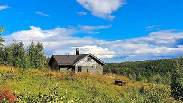 A rustic cabin with a sloped roof, surrounded by dense forest and under a clear blue sky.