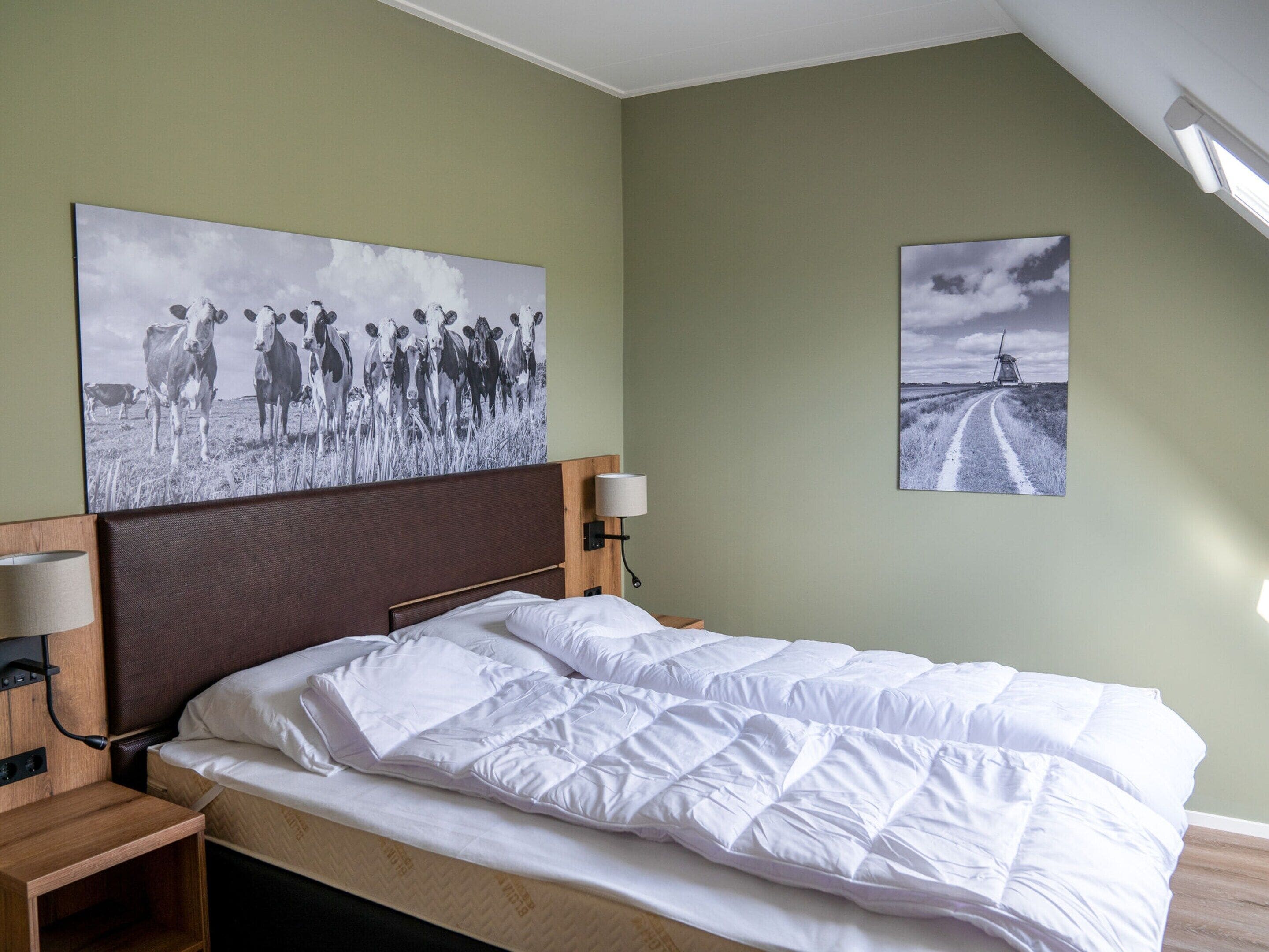 A neatly made bed with white bedding, a wooden headboard, a bedside table with a lamp, and two wall-mounted pictures depicting cows and a windmill.