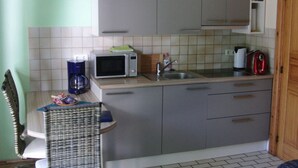 A small kitchenette with a microwave, coffee maker, and toaster. There is a wicker chair and a wooden door. - Karlo 4 Modern Retreat (Büsum)