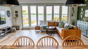 A spacious living room with a large wooden dining table, comfortable seating, and a view of a lake through large windows. - Oceanhouse Comfortable Holiday Residence (Wieringerwerf)