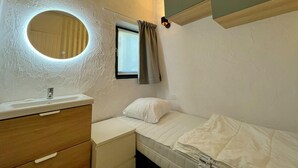 A small, compact room with a single bed, a bedside table, a mirror, and a window with curtains.