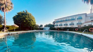 Una piscina con agua azul cristalina, rodeada por un edificio blanco con balcones y una palmera.