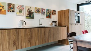 A modern kitchen with wooden cabinetry, a central island, a sink, and a coffee machine. There are six floral paintings on the wall above the counter. - Holiday Home for Family (Ouddorp)
