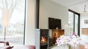 A modern living room with a wood-burning stove, a comfortable leather sofa, a wooden table, and a vase of flowers. - Holiday Home for Family (Ouddorp)