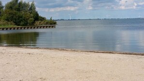 A calm lake with a sandy shore, a wooden dock, and wind turbines in the distance. - Holiday Home Deichblick 58 (Andijk)
