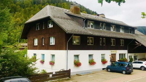 Ein zweistöckiges Gebäude mit geneigtem Dach, weißen Wänden und Holzverzierungen. Blumenkästen befinden sich vor den Fenstern im unteren Bereich und ein Auto steht auf der Auffahrt.