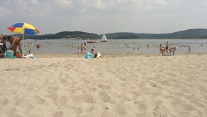 Een zandstrand met mensen die zwemmen en zonnebaden, een zeilboot in de verte, en een kleurrijke parasol.