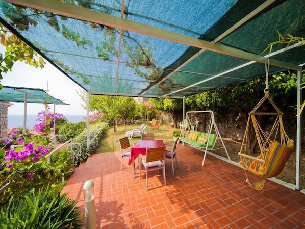Relax in the covered patio area with a table, chairs, swing, and hanging chair, surrounded by a garden with beautiful sea views.