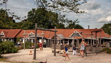 Een zandrijk buitengebied met houten speeltoestellen en een gebouw met een dak van rode tegels.