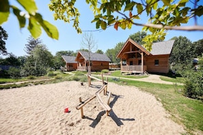 A wooden cabin with a deck, surrounded by a sandy area and greenery. - Holiday Home in Brabant With Private Spa (Uden)