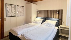 Una cama bien tendida en una habitación con cabecera de listones, dos cuadros enmarcados en la pared y una mesita de noche con lámpara.