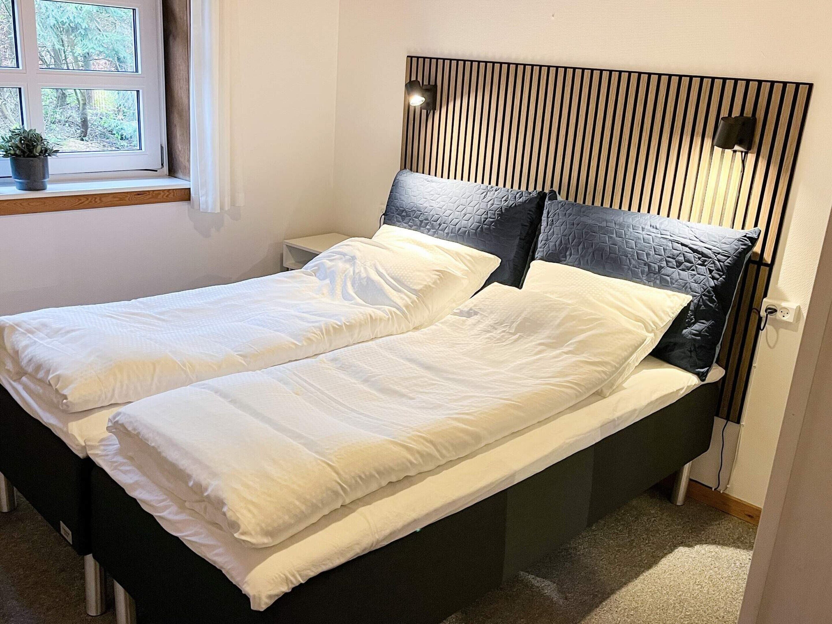 A double bed with white bedding, a wooden headboard, a bedside table, and a window with a view of trees.