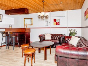 A room with a wooden ceiling, a round table, a leather sofa, two wooden stools, a bar area with a mirror, and wall-mounted artwork. - 4 Star Holiday Home in Oster Assels (Øster Assels)
