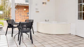 Een badkamer met een bad, een wastafel en een toilet. Er is een raam met uitzicht op een bakstenen muur en een deur die leidt naar een buitenruimte met stoelen en een tafel.