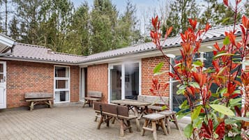 Een bakstenen gebouw met een pannendak, een houten tafel en banken, en een tuin met bomen en struiken.