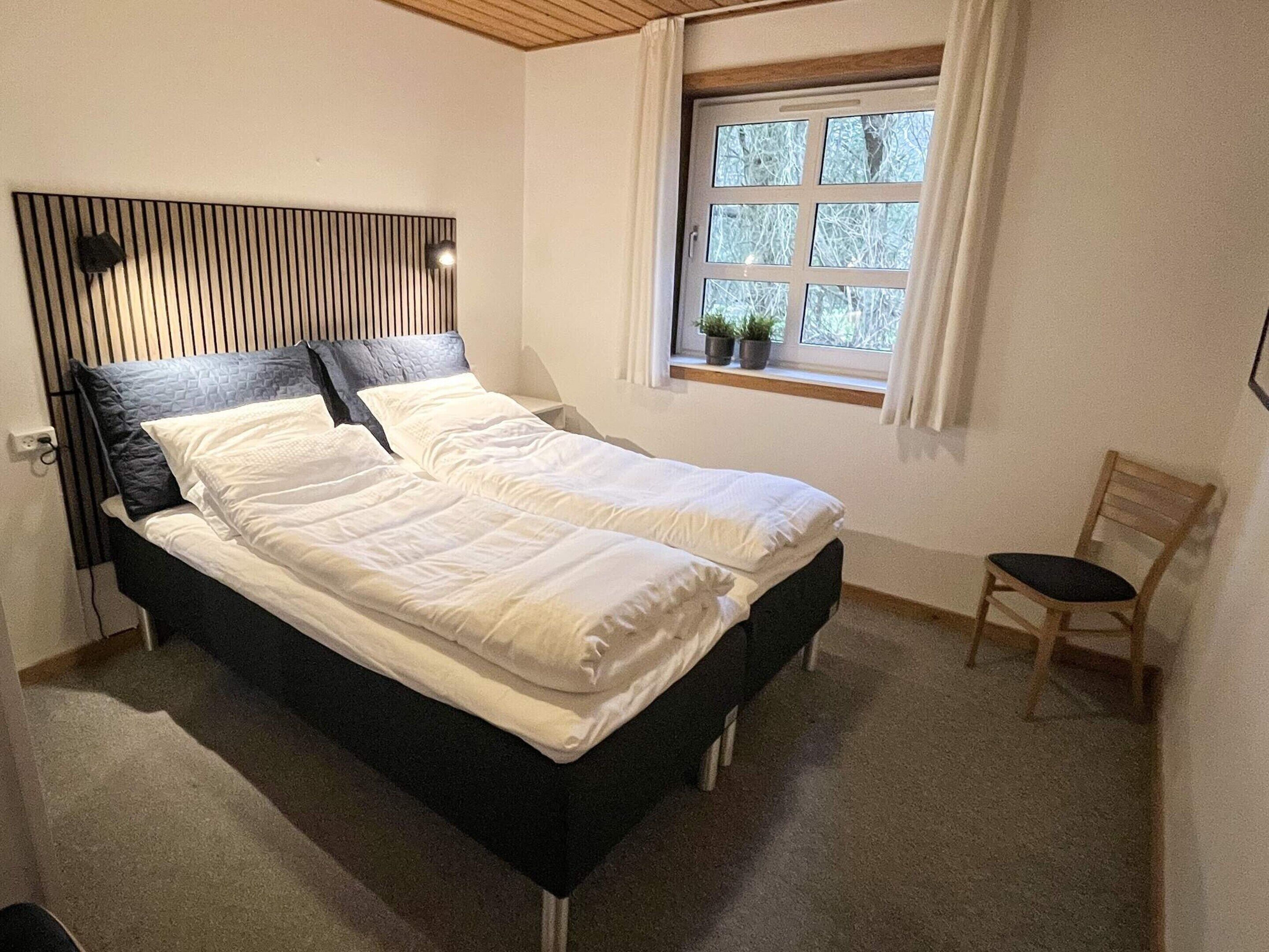 Ein Schlafzimmer mit einem Bett, einem Fenster mit Vorhängen, einem Stuhl und einer Wandleuchte.