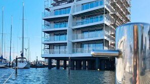 Un bâtiment moderne doté de nombreux balcons et de grandes fenêtres, situé au bord d’un plan d’eau où se trouvent des bateaux.