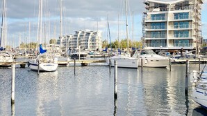 Un port de plaisance où sont amarrés de nombreux bateaux, un bâtiment moderne doté de balcons et un ciel dégagé.