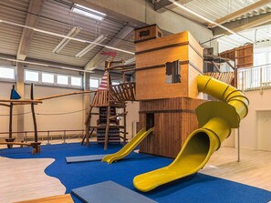 Een binnenspeelruimte met een glijbaan, klimconstructies en een houten speelhuis.