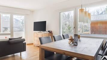 Uma sala de estar moderna com uma mesa de jantar de madeira, cadeiras pretas, um sofá e uma TV em um armário de madeira. Janelas amplas oferecem vista para o exterior.