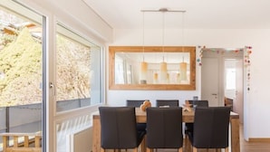 Un coin repas avec une table en bois et des chaises noires, un grand miroir au-dessus de la table et une vue sur les arbres à l’extérieur.