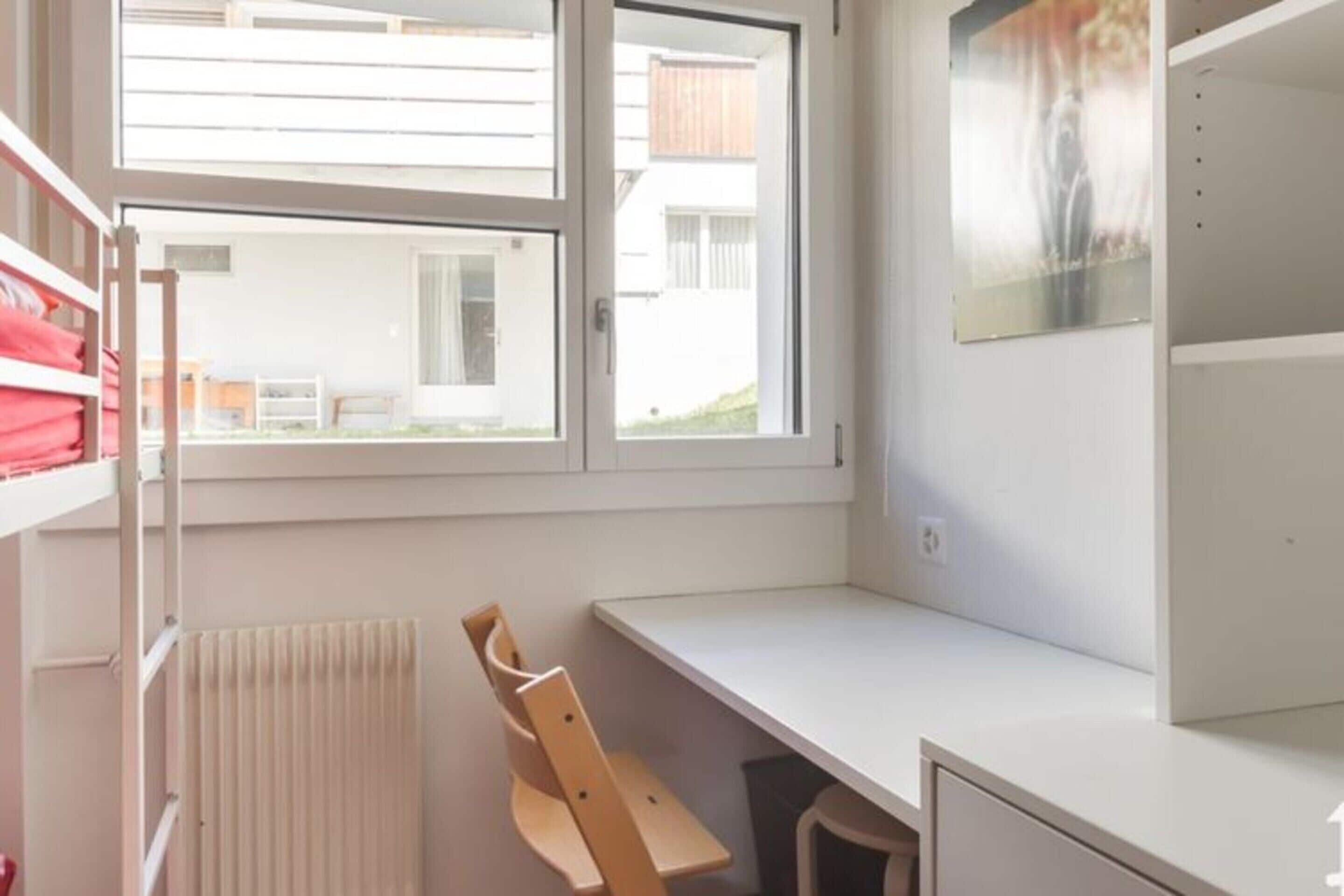 Una habitación con un escritorio blanco, un radiador, una ventana y una silla.
