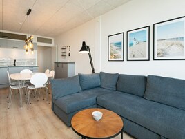 En moderne stue med en grå sofa, et rundt træbord og et spiseområde med hvide stole og et glasbord.