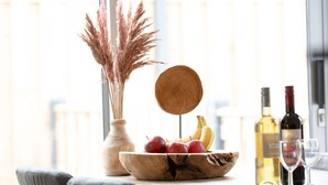 Une table à manger sur laquelle se trouve un saladier en bois rempli de fruits, un vase contenant des plantes séchées et des bouteilles de vin.