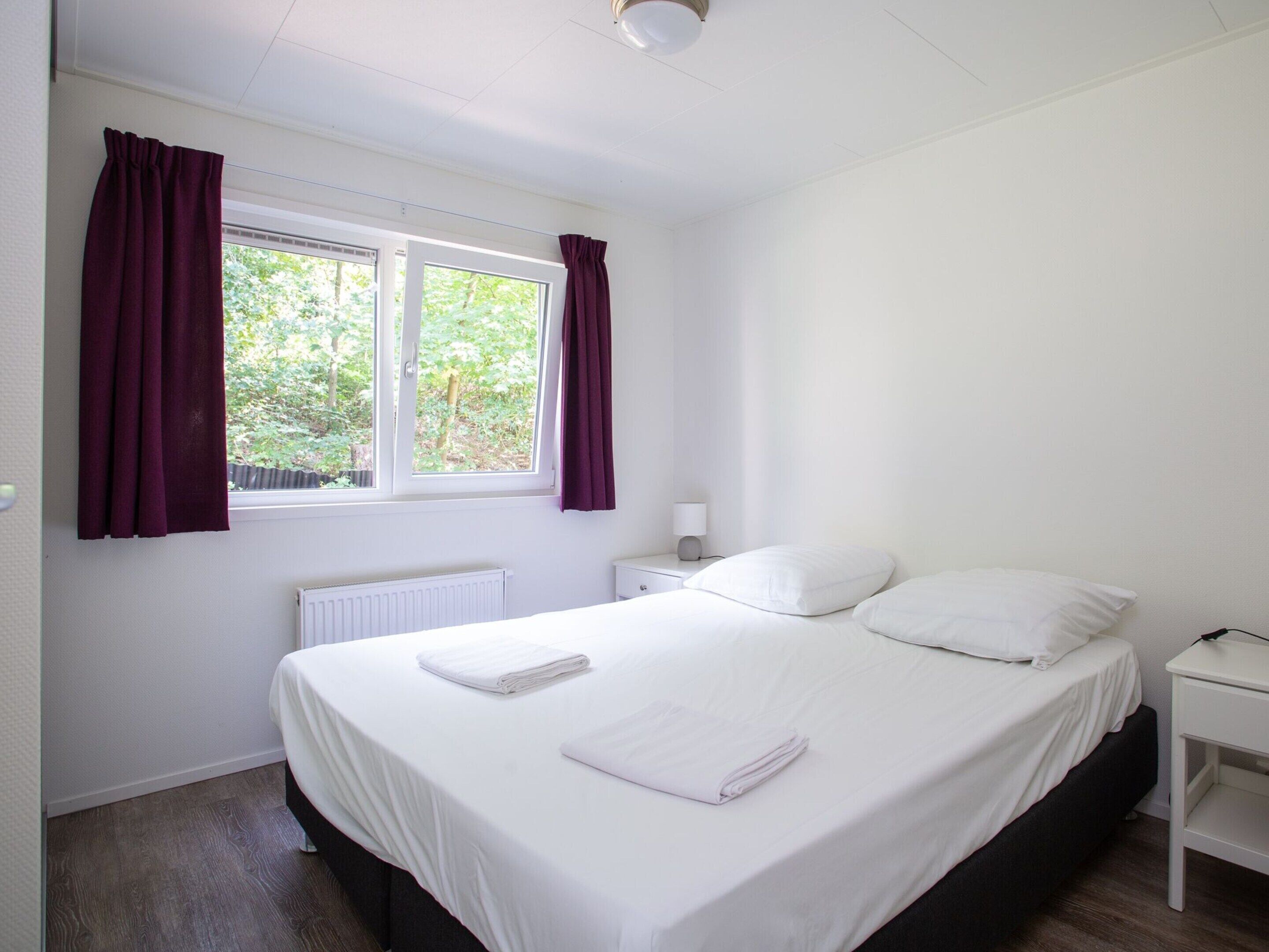 Une chambre à coucher avec un lit, des rideaux blancs, une fenêtre donnant sur un paysage verdoyant, une table de chevet avec une lampe et un radiateur.