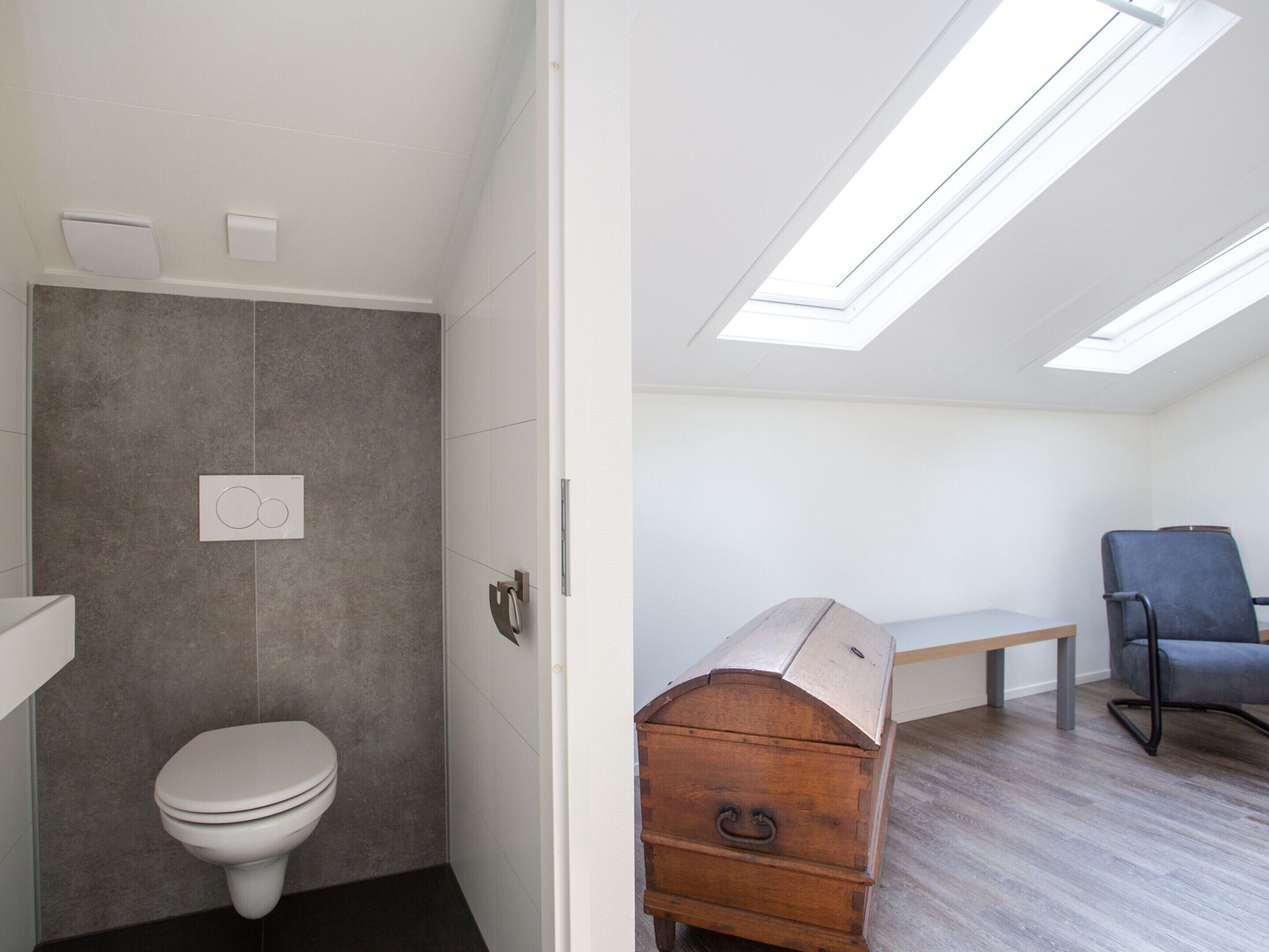 Une petite pièce comprenant des toilettes, une commode en bois, un banc et une chaise.