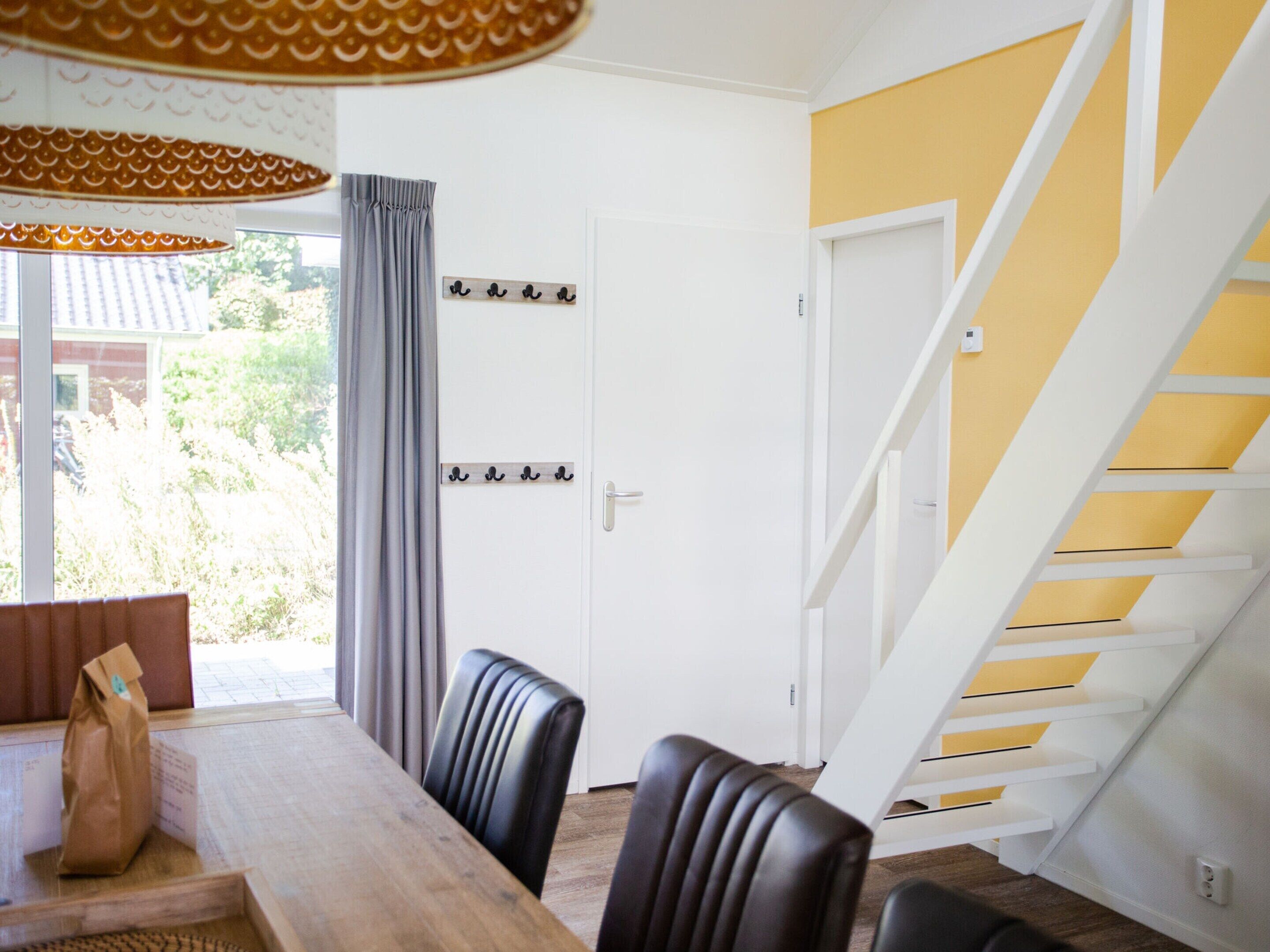 A dining area with a wooden table and black chairs, a white door with a coat rack, and a staircase with yellow accents.