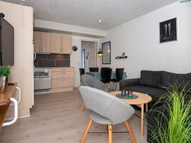 Un espacio de estar moderno con un sofá gris, una pequeña mesa redonda y una zona de cocina con armarios de madera y un horno empotrado.