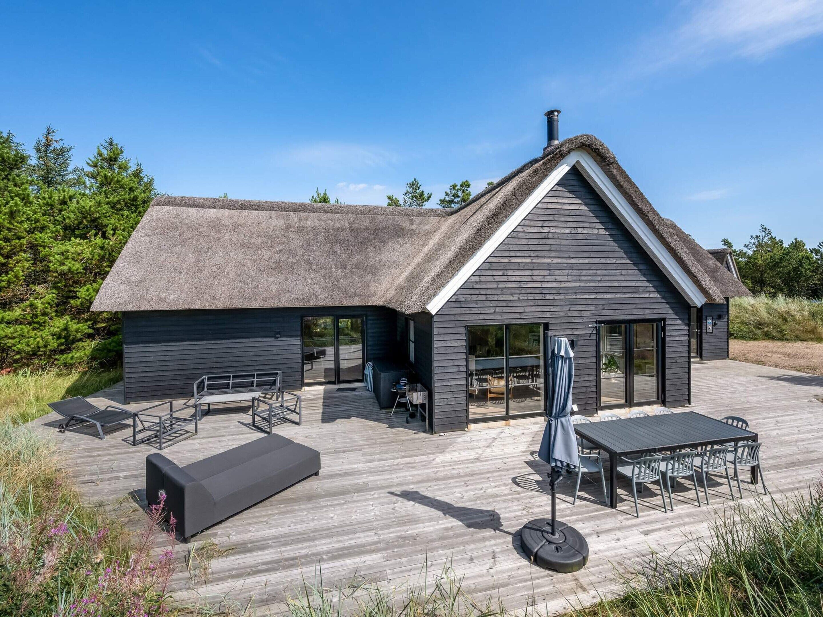 A modern house with a thatched roof, large windows, and a wooden deck with outdoor furniture.