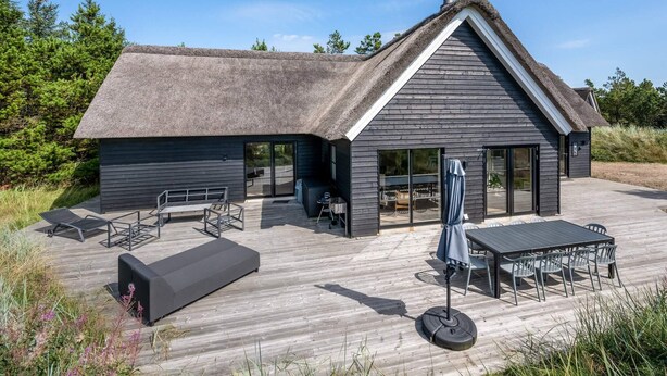 Et moderne hus med stråtækt tag, store vinduer og en træterrasse med udendørs møbler.