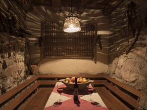 Una bodega rústica con paredes de piedra, un banco de madera, una mesa con un cuenco de fruta y copas de vino, y un candelabro colgante.