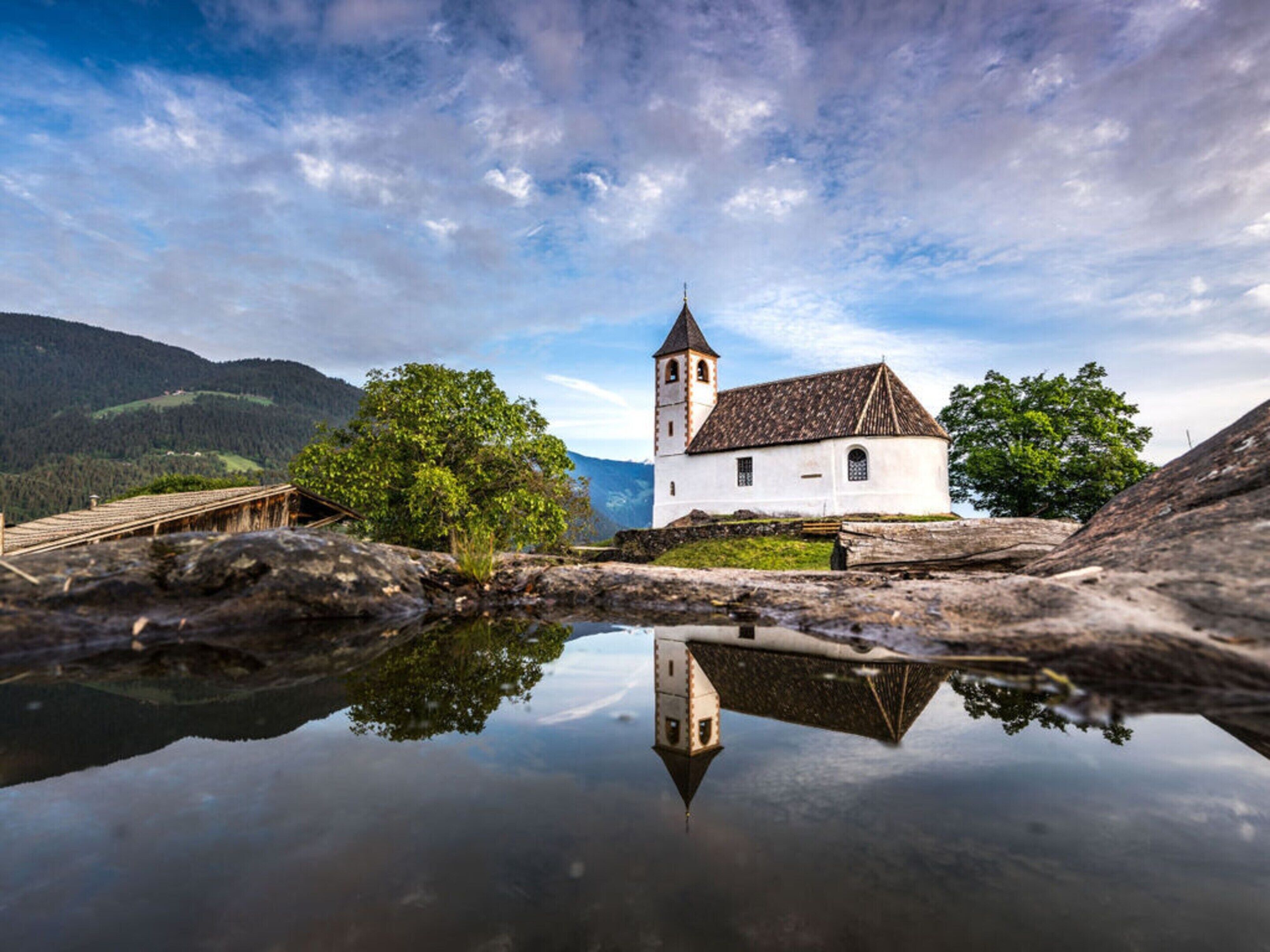 Una iglesia blanca con un campanario, rodeada de árboles y montañas, reflejada en un cuerpo de agua tranquilo.