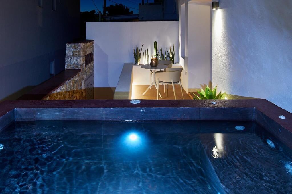 A swimming pool with a stone edge, a small table with chairs, and potted plants.
