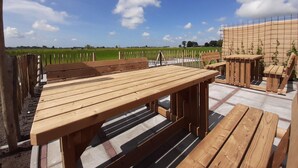 Un patio al aire libre con bancos y mesas de madera, vista a un campo verde y un cielo azul despejado.