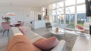 A modern living room with a sofa, a coffee table, and a TV. There is a dining area with a table and chairs. The room has large windows and a kitchen in the background. - Dyke-view Stay With Terrace (Wemeldinge)