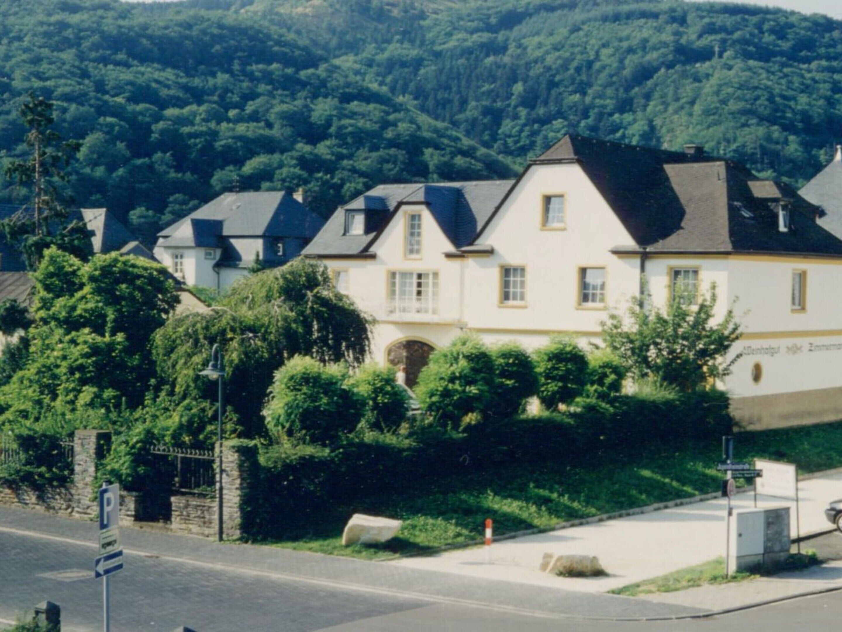 A row of white houses with dark roofs, surrounded by lush greenery and a forested hillside.