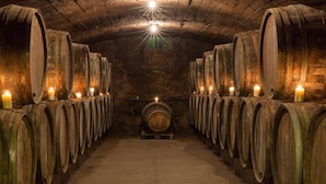 Uma adega com barris de madeira dispostos em fileiras, iluminada por velas, e um barril central ao fundo.