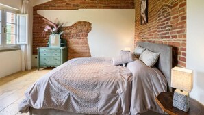 A bedroom with a brick wall, a wooden ceiling, a bed with a grey headboard, and a nightstand with a lamp. - Villa al Molino Comfortable Holiday Residence (Fauglia)