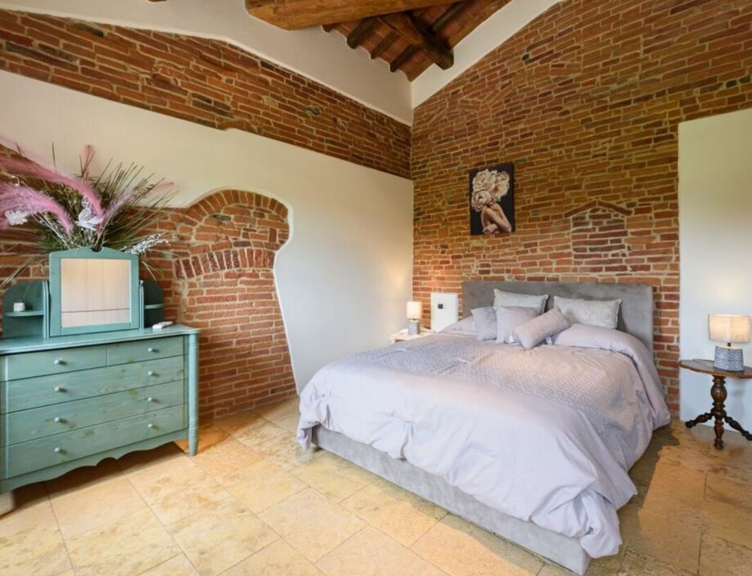 Une chambre avec un mur en briques, un lit avec une literie grise, une table de chevet avec une lampe et une commode ornée d’un vase de fleurs.