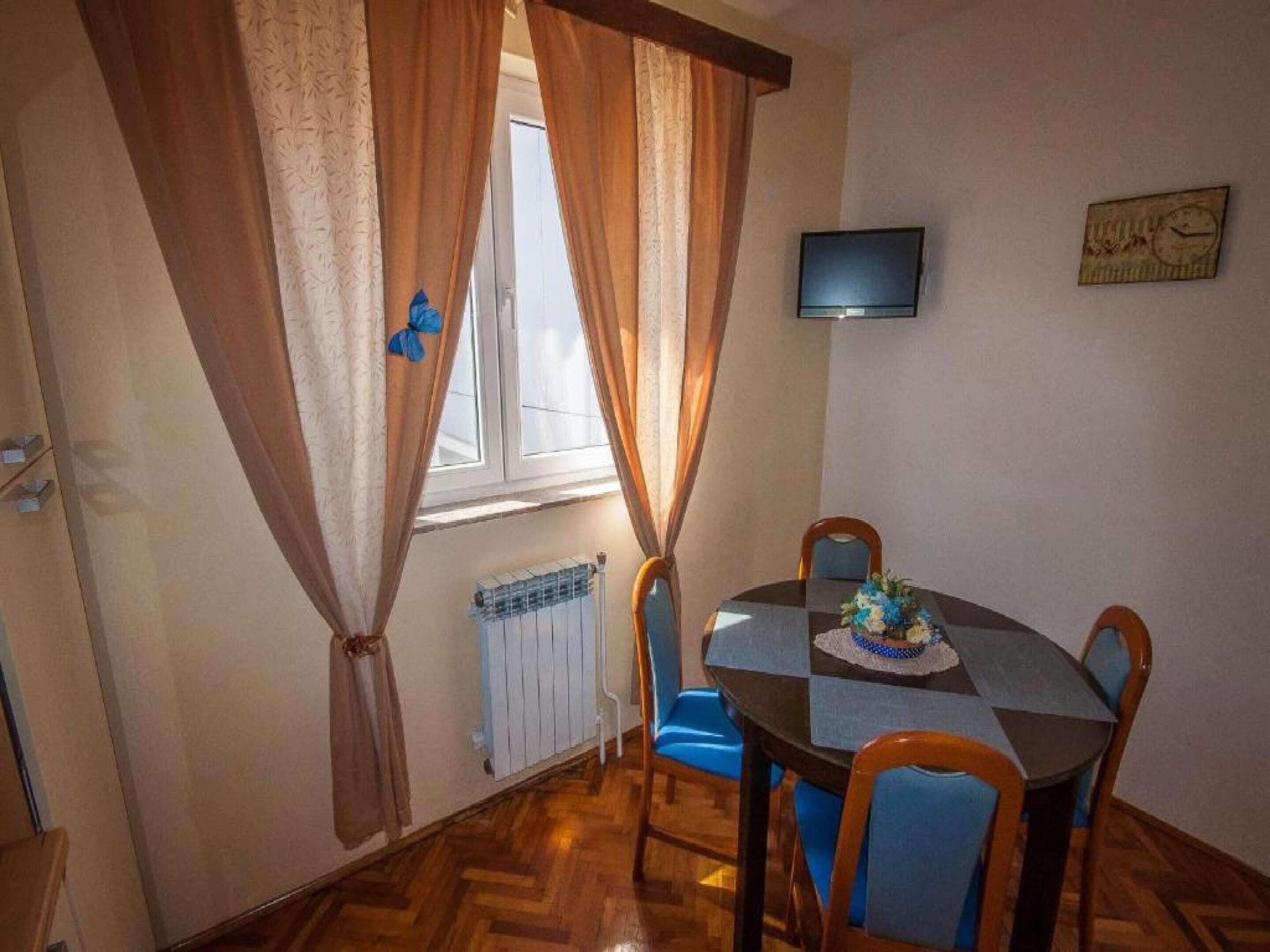 A dining area with a round table, blue chairs, a flower centerpiece, a TV on the wall, and a window with curtains.