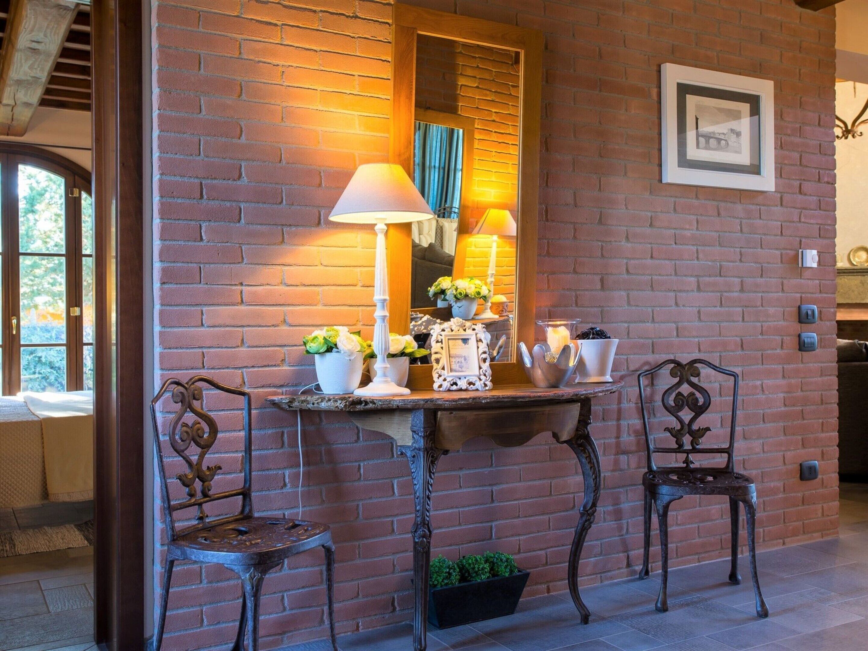 A brick wall with a mirror, a table with decorative items, and two ornate chairs.